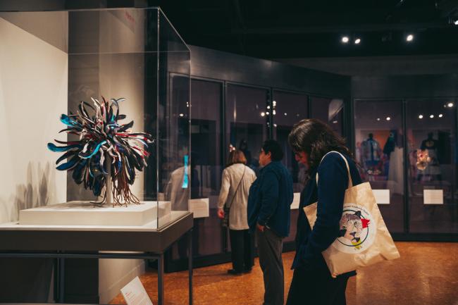 People are viewing various exhibits in a dimly lit art museum. A sculpture that appears like an abstract burst of colored materials is in a glass case to the left. Visitors are observing the displays, and one person carries a tote bag with a colorful design.