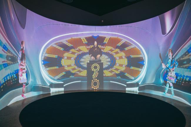 A futuristic room features a DJ at a console with a large "315" displayed. The background is filled with colorful, dynamic patterns. Two individuals in vibrant costumes are on either side, appearing to perform or entertain amidst the visual spectacle.