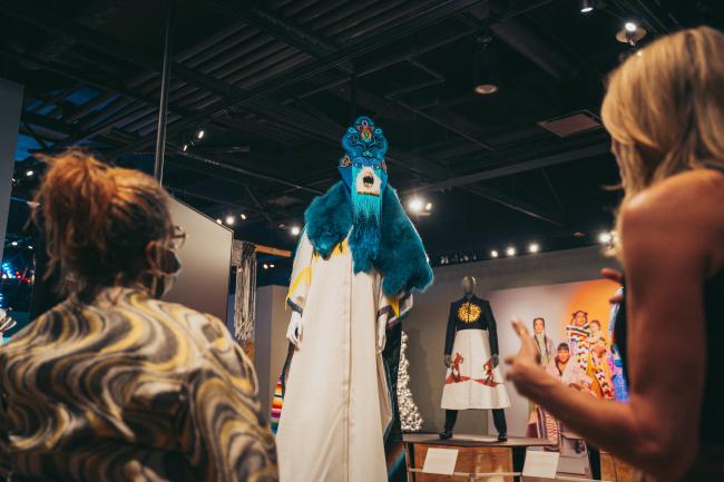 Two people observe an elaborate costume on display in a dimly lit exhibition. The costume features vibrant blue fur and an intricate mask with a headdress. Another costume on a mannequin and various colorful outfits are visible in the background.