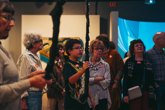 A diverse group of people of various ages stand attentively in an indoor setting, possibly a museum or gallery. The focus is on a young boy holding a long, dark object while others look on with interest. The background features framed artwork and a blue-lit display.