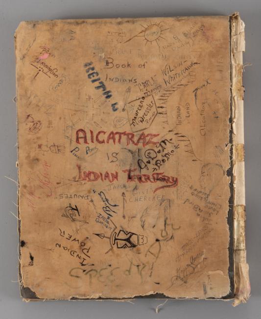 An old book cover, worn and frayed, with various handwritten messages and doodles. Prominent text reads, "Alcatraz is Indian Land," surrounded by names and other writings. The cover is beige and shows signs of heavy use.