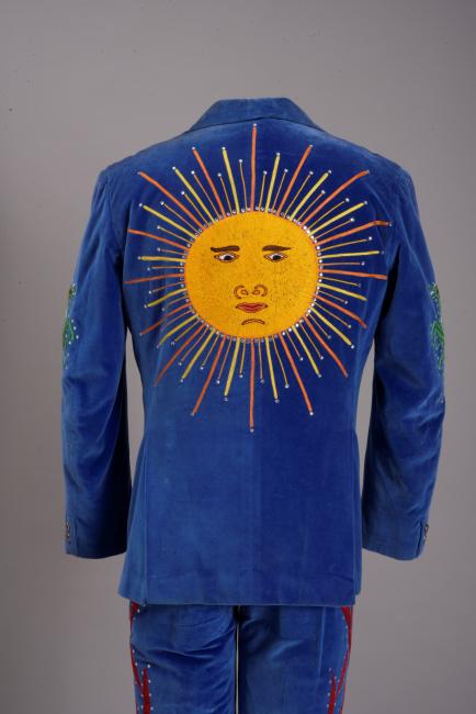 A blue velvet suit jacket with an embroidered yellow sun featuring a face on the back. The sun has rays extending outward, and the jacket has intricate designs on the sleeves. The matching pants have decorative patterns.