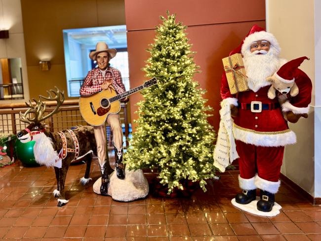 Christmas display featuring a cowboy with a guitar, a reindeer wearing decorations, a lit Christmas tree, and a Santa Claus figure holding a teddy bear and a gift. The scene is set indoors with festive decor.