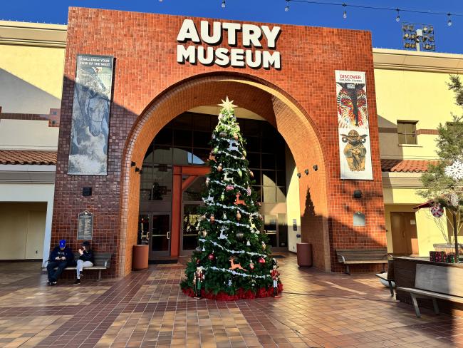 A large Christmas tree adorned with decorations stands in front of the entrance to the Autry Museum. The building features an archway with "Autry Museum" above it and banners on either side. Two people are sitting on benches nearby.