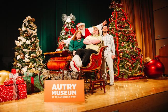 A group of four people pose in a festive setting at the Autry Museum. Santa sits in a sleigh surrounded by decorated Christmas trees and wrapped gifts. A sign reads "Autry Museum of the American West" in front of them.