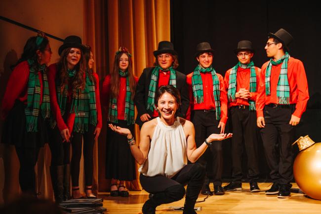 A group of performers in festive attire, including red shirts, green plaid scarves, and black hats, stand on a stage. A woman in front is kneeling, smiling with her arms open wide. Curtains and stage decorations are visible in the background.