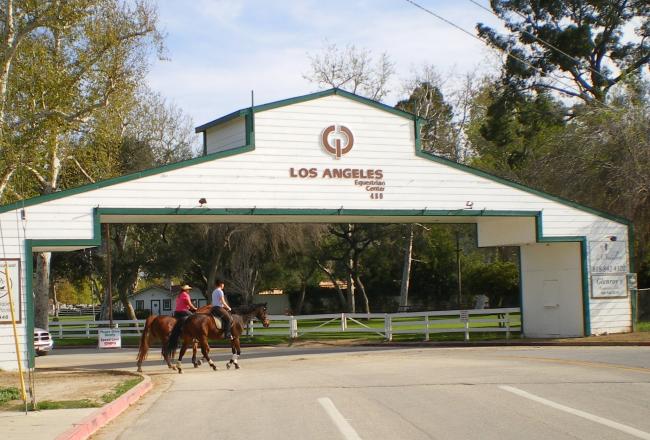 Two people ride horses under a large white wooden archway labeled "Los Angeles Equestrian Center" on a sunny day, with trees and a fenced green area in the background.