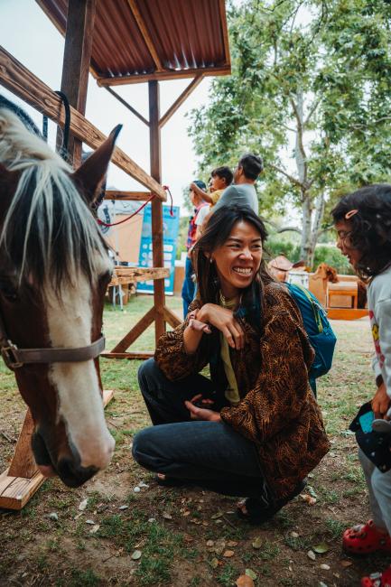 A woman crouches and smiles at a young girl near a horse at an outdoor event, with trees and other people visible in the background.