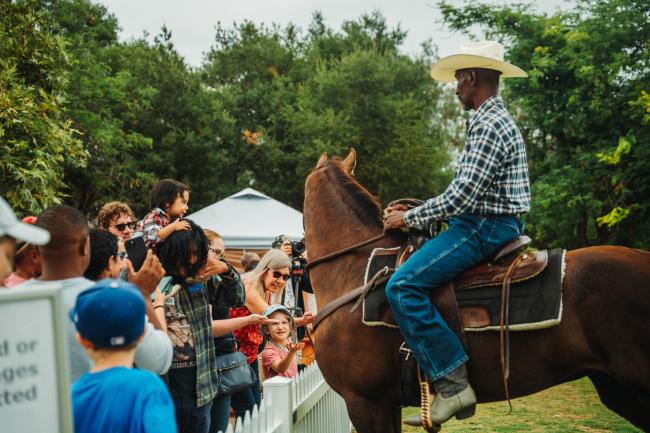 A man in a cowboy hat and plaid shirt sits on a brown horse while a group of children and adults gather near a white fence to greet him and take photos, surrounded by trees outdoors.