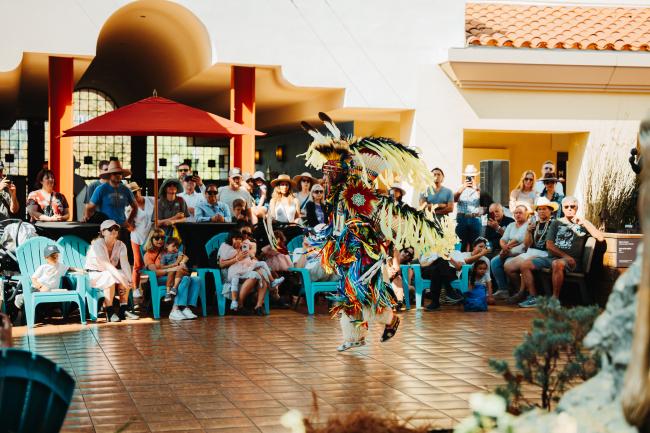 A Native American dancer in vibrant traditional regalia performs outdoors before a seated crowd, with colorful feathers and ribbons swirling as people watch and take photos on a sunny day.