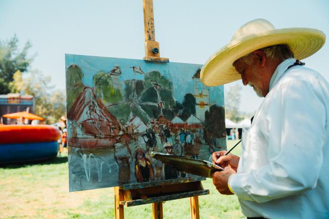 An older man in a wide-brimmed hat paints an outdoor festival scene on a canvas set on an easel, surrounded by grass and tents on a sunny day.