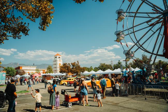People enjoy a lively outdoor fair with a Ferris wheel, classic cars, vendor tents, and trees under a sunny blue sky. Families and children walk around, and a clock tower is visible in the background.