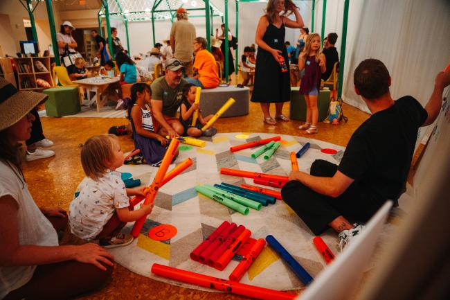 Children and adults sit on a rug in a brightly lit playroom, playing with colorful plastic tubes. Other groups are engaged in activities in the background, creating a lively, interactive environment.
