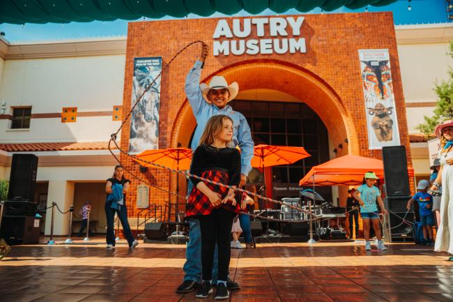 A man in a cowboy hat twirls a lasso above a young girl’s head outside the Autry Museum, with people and red umbrellas in the background on a sunny day.