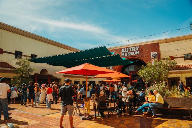 A crowd of people gather outside the entrance of the Autry Museum on a sunny day. Some sit under red umbrellas at tables, while others stand or walk in the courtyard, which is decorated with greenery and string lights.