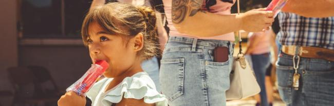 A young girl with brown hair eats a red popsicle outdoors, standing next to adults who are also holding popsicles. The scene appears sunny, and the people are casually dressed.