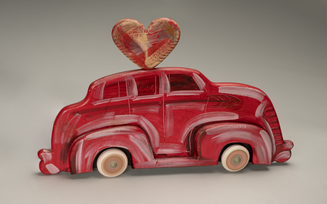 A toy car painted red with white accents, featuring a large red and gold heart shape attached to its roof, set against a plain gray background.