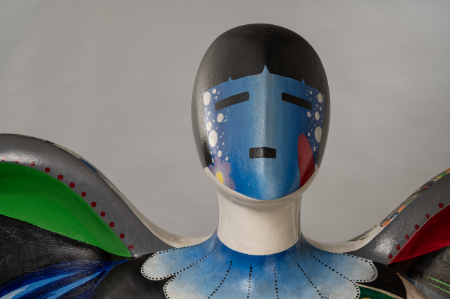 A close-up of a colorful abstract sculpture featuring a humanoid figure with a smooth face painted blue and white, dark hair, and vivid patterns on its wings and chest, set against a plain gray background.