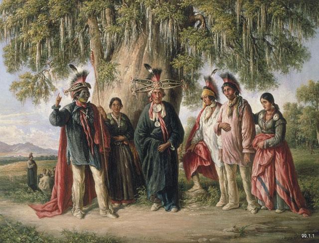 Close up of Six Kickapoo Indians, Chief and Family without its frame. The oil painting shows a group of 6 people gathered outside, in the foreground, with a small cluster of individuals in the background, behind a tree. The foreground group are ornately dressed with several people wearing headdresses. The person on the most left has green face paint and a large staff. They are gesturing with their hand, looking towards the person in the middle, that faces out towards the viewer.