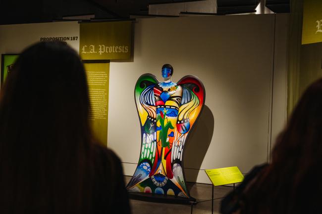 A colorful angel sculpture with abstract patterns is displayed in a museum exhibit about L.A. protests, with two people viewing it from the foreground.