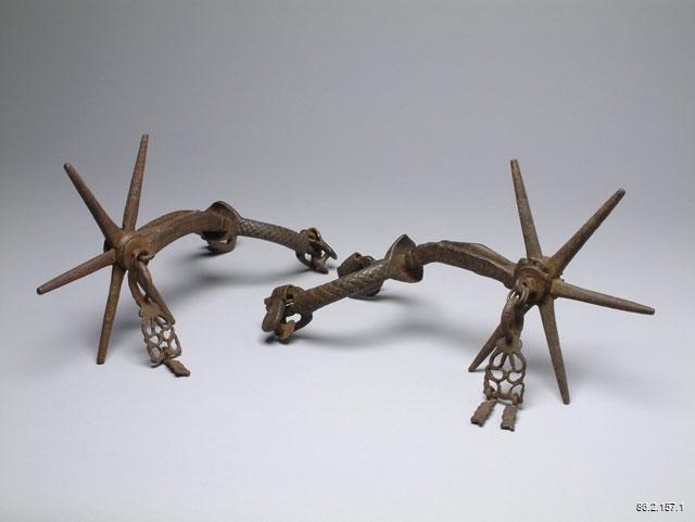 Two rusted, metal spurs propped up by their heel bands and rowels. Neither spur has a strap. The heel band has a braided rope texture. The rowels have 6, widely spaced, conical prongs. Attached to the rowel pin covers are rusted metal ornaments. The ornaments have a symmetrical cut-out pattern with a small, square rectangle attached to the bottom.