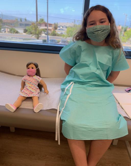 a young girl is masked and dressed for a medical visit, a doll to the left also wears a mask