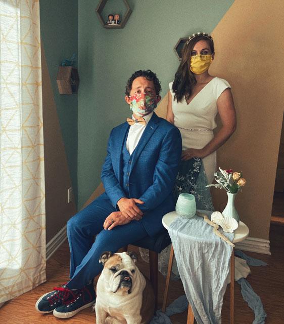 A wedding portrait of a heterosexual couple wearing masks, a dog is at their feet