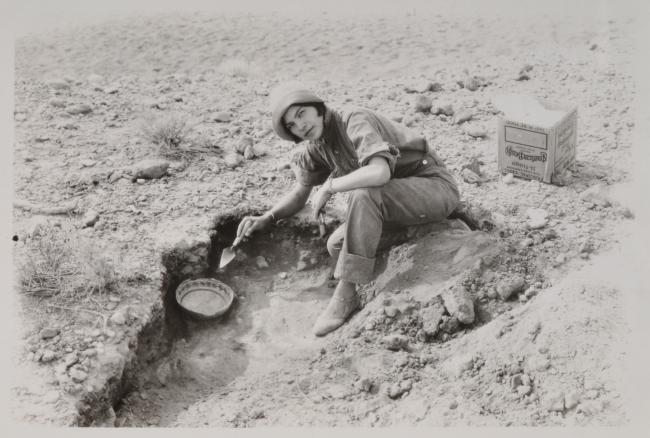 woman sits at archaeological dig, a basket is in the trench