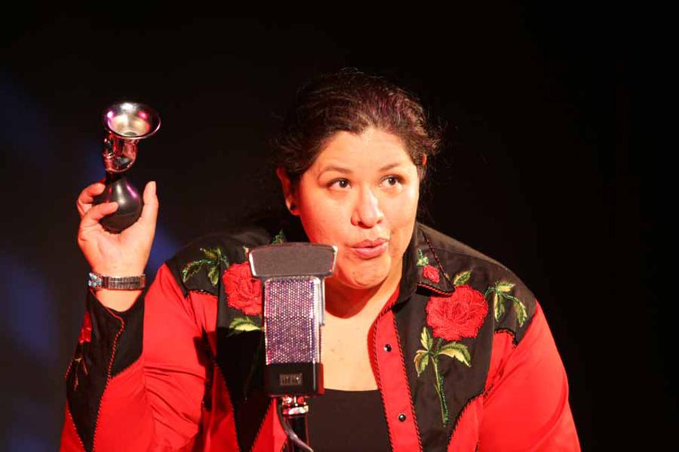 woman speaks at a microphone wearing a red embroidered shirt and holding a metal object