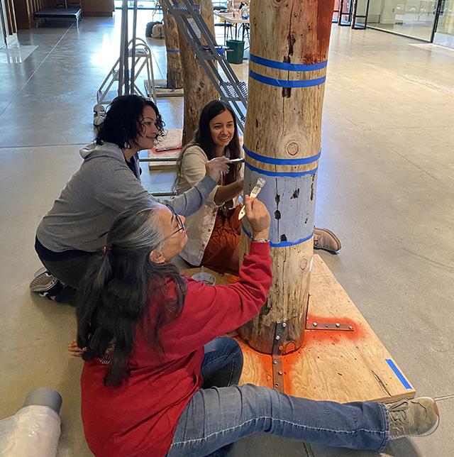three women sit on the floor painting a design on a wooden pole