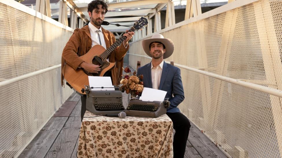 Two men standing on an outdoor bridge with a netted railing. One stands playing an acoustic guitar, wearing a brown suede jacket with fringe. The other sits at a table covered with a floral cloth, wearing a white hat and suit, with two typewriters in front of him.