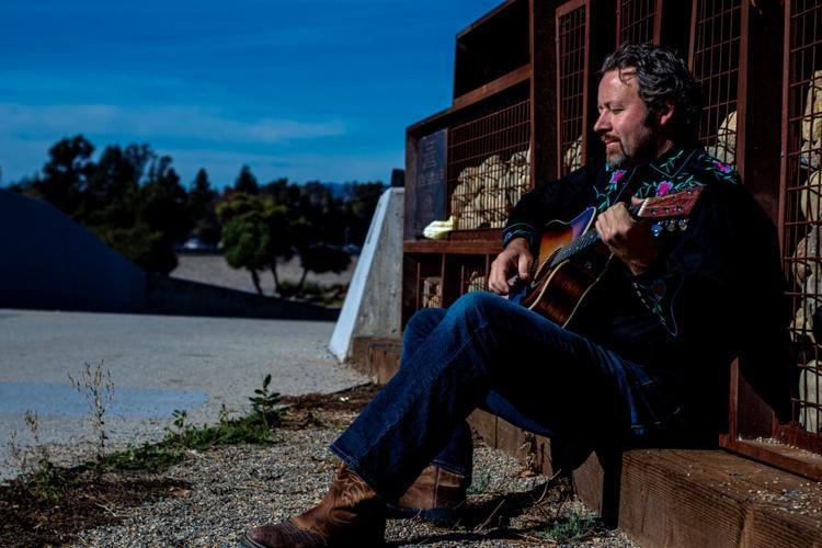 A man sits on wooden steps outdoors, playing an acoustic guitar. He's wearing jeans, cowboy boots, and a black shirt with colorful embroidery. The background features trees and a partially cloudy blue sky.