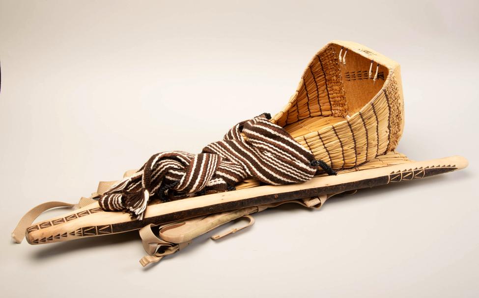 A traditional woven wooden cradleboard with a curved hood and a brown and white striped cloth wrapped around it, used for carrying infants. The design features straps and carved decorative patterns.