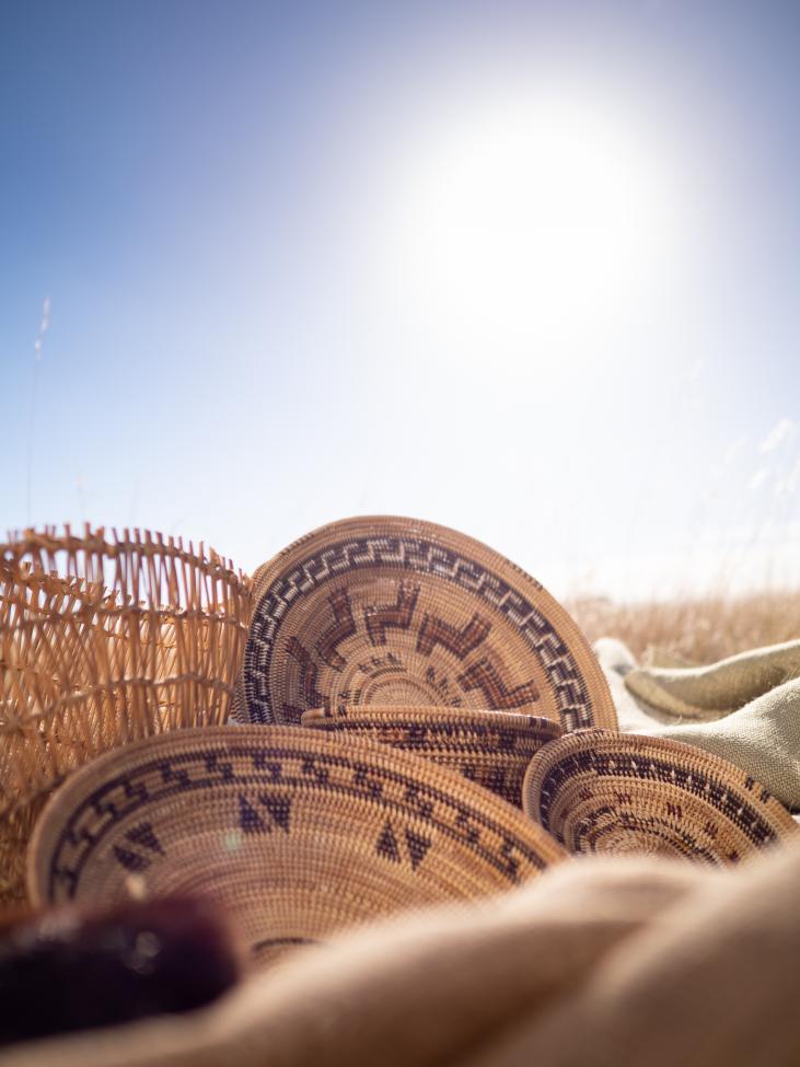 Woven baskets with intricate patterns rest on a blanket in a sunlit field, with tall grass and a bright sky in the background.