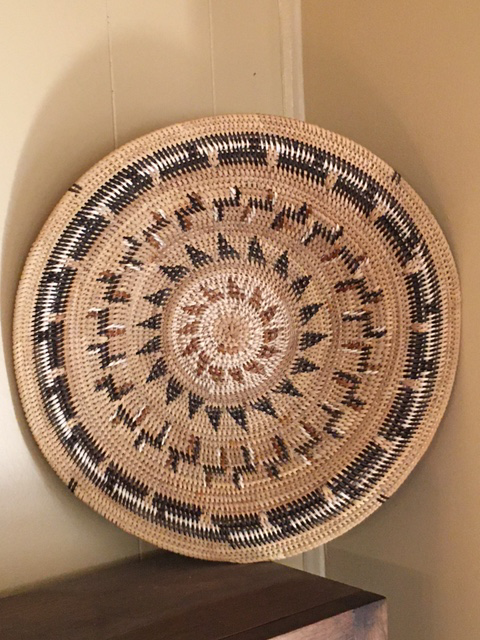 A large, round woven basket with intricate geometric patterns in natural, black, and brown colors is displayed upright against a beige wall on top of a dark wooden surface.