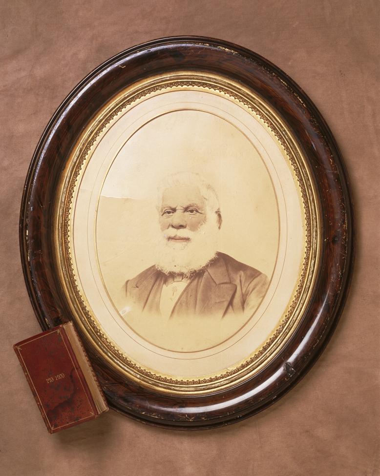 Oval-framed sepia portrait of an older man with a full white beard, wearing a suit. A red book titled "The Negro" rests against the frame on a brown surface.