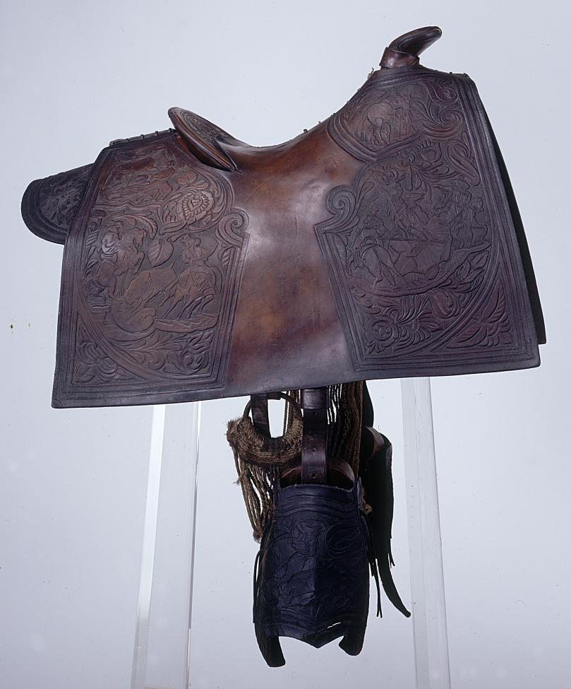 An ornate dark leather horse saddle with intricate carvings featuring detailed scenes and patterns, displayed on a stand against a plain white background.