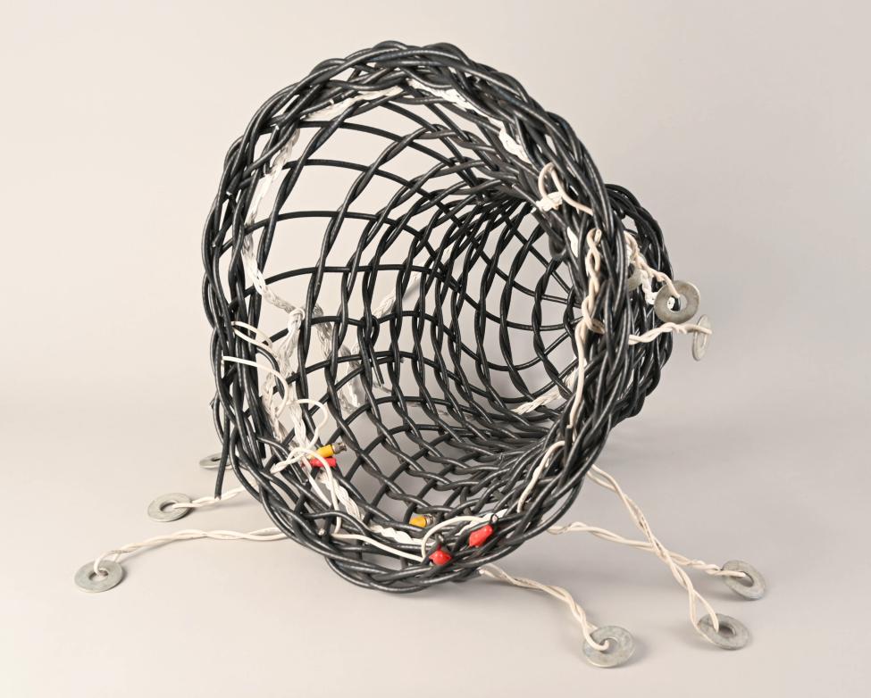 A black, cylindrical, woven fishing trap with white string and metal weights attached, displayed on a light gray background. The trap is open at one end and tapers slightly at the other.