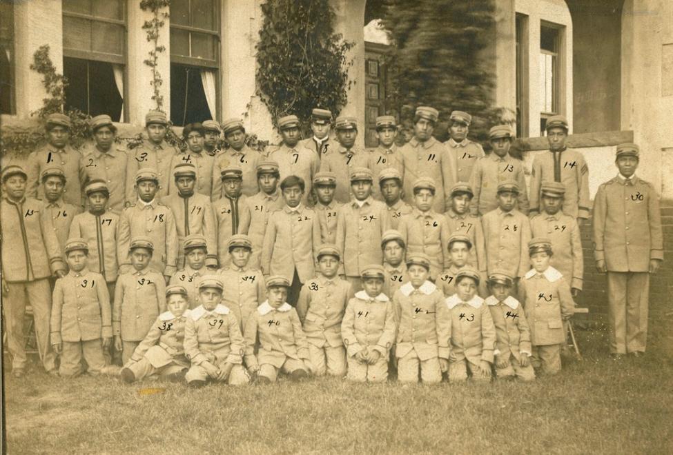 A sepia-toned photo of a large group of boys and young men in uniform and caps, standing and sitting in rows outside a building. Numbers are handwritten on each individual's uniform for identification.