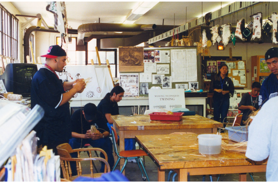 A group of people sit and stand around tables in a classroom, weaving and working with natural materials. Artworks and posters decorate the walls, and supplies are scattered on the tables.
