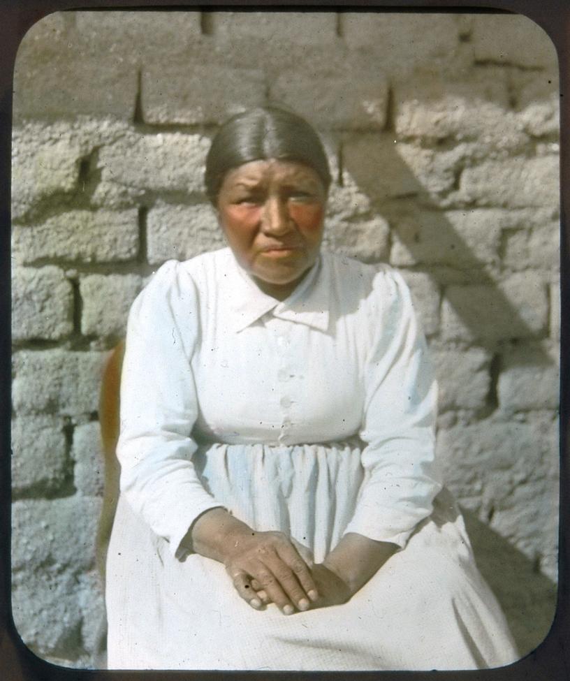 A woman with dark hair parted in the middle and tied back sits against a brick wall, wearing a long-sleeved white dress and looking at the camera with her hands folded in her lap.