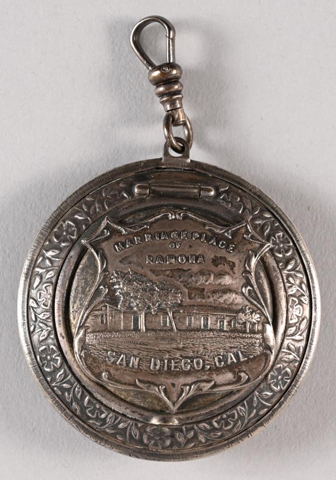 A round silver medallion with an engraved image of a house and trees, inscribed with "Marriage Place of Ramona" and "San Diego, Cal," with decorative patterns around the border and a loop at the top for attachment.