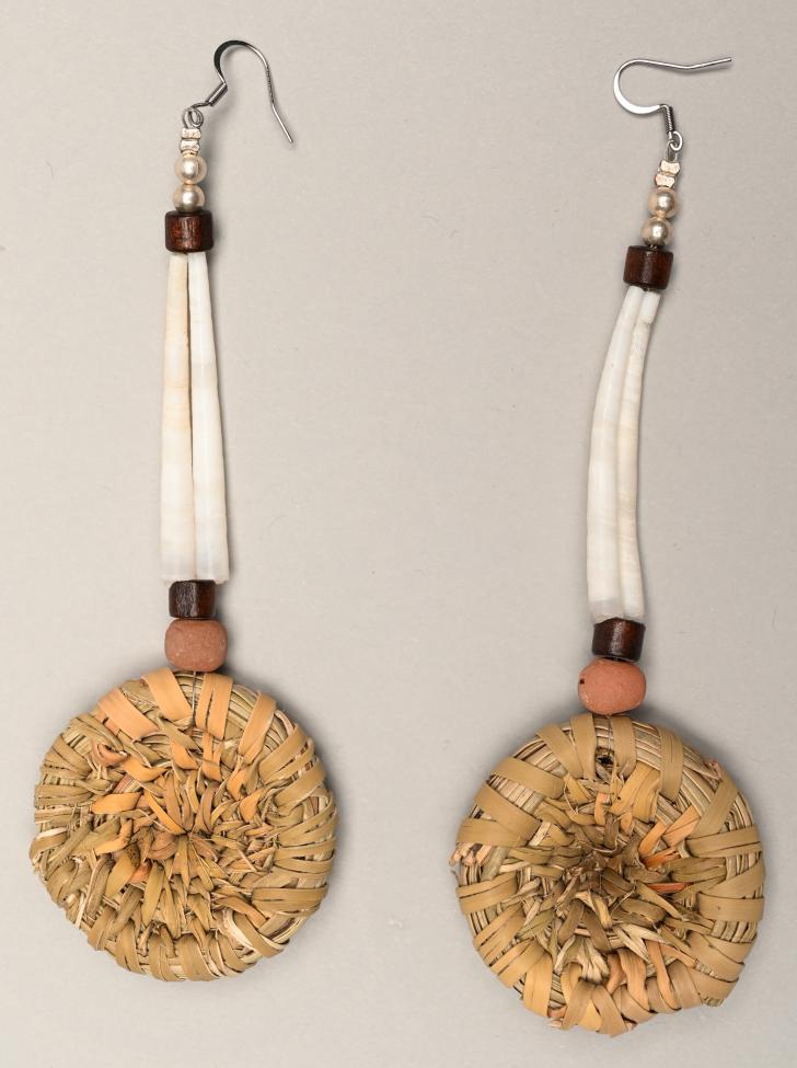 A pair of handmade earrings featuring round, woven straw discs, brown and pink beads, and elongated white beads, all hanging from metal hooks on a neutral background.