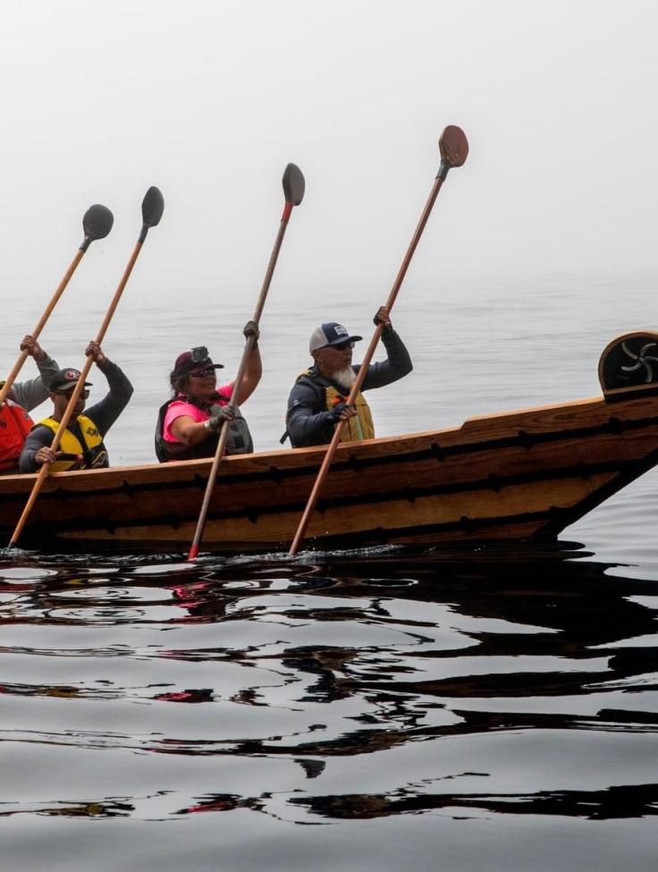 Four people paddle a wooden canoe on calm, misty water, each wearing life jackets and hats. The sky and background are foggy, creating a serene and peaceful atmosphere.