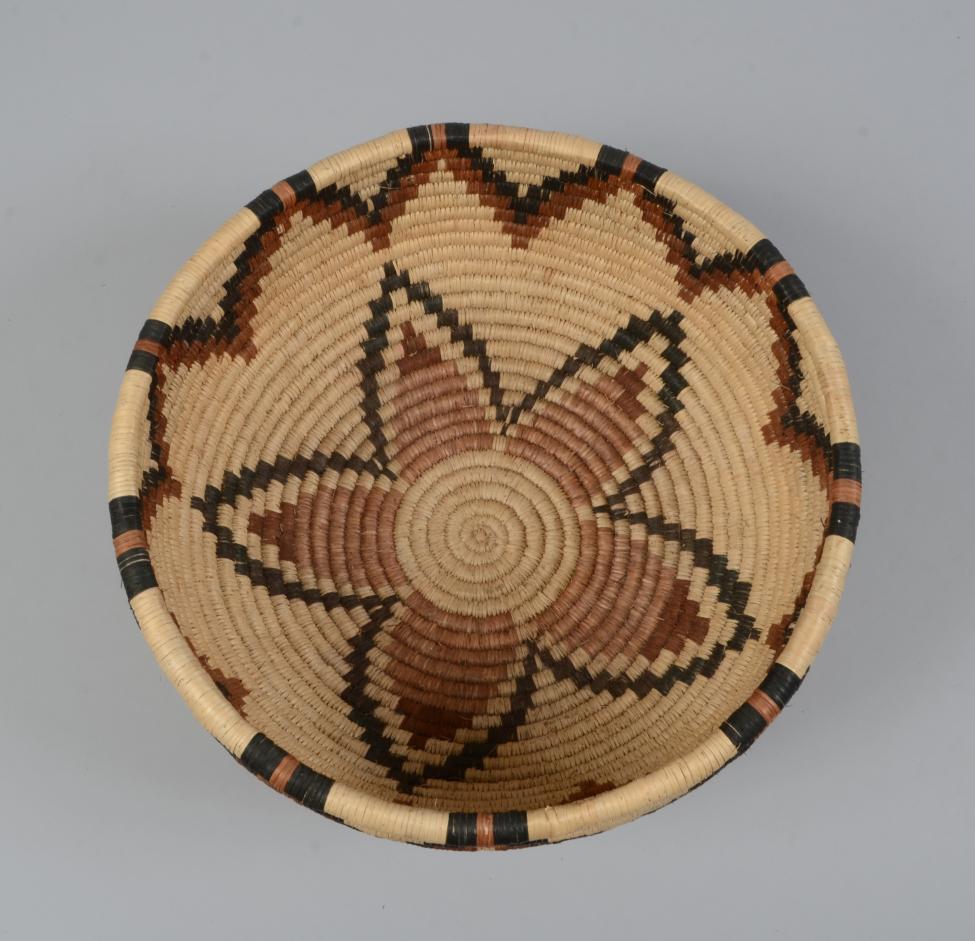 A woven basket with a beige and brown zigzag border and a central star or flower pattern in shades of brown and black on the interior, viewed from above against a plain background.