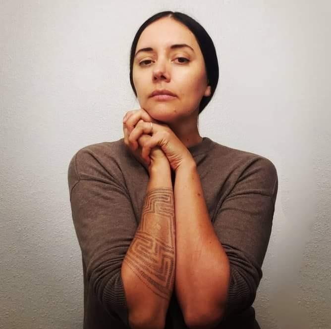 A woman with dark hair pulled back gazes at the camera, hands clasped under her chin. She wears a brown long-sleeve top and has a geometric tattoo on her left forearm. The background is plain and light-colored.