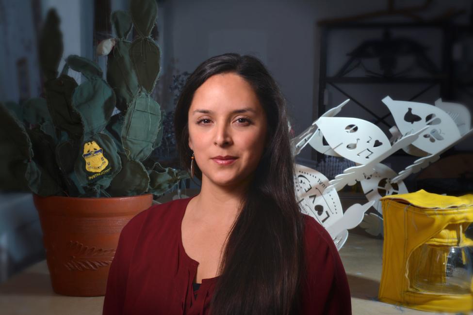 A woman with long dark hair wearing a maroon top stands indoors. Behind her are a potted cactus, a yellow object, and a white paper-like sculpture with cutout designs.
