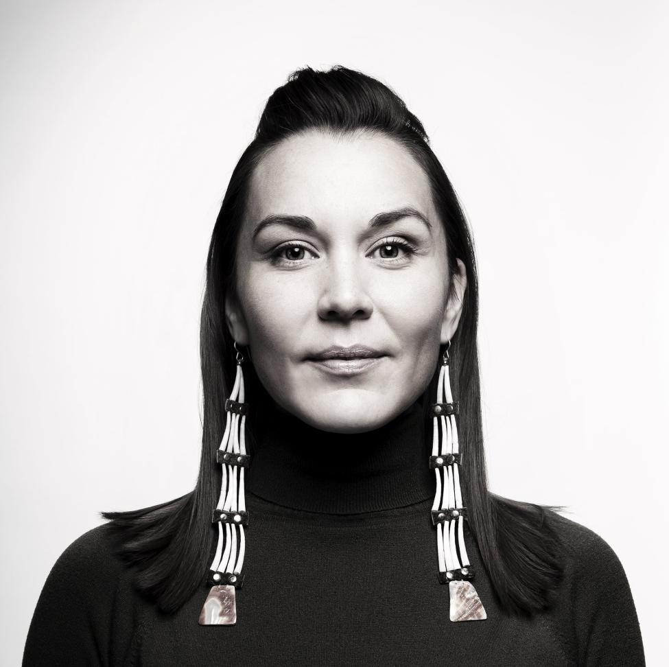 A woman with straight, shoulder-length dark hair wears a black turtleneck and long, distinctive earrings made of white and dark segments with pinkish stone pendants. She looks calmly at the camera against a plain background.