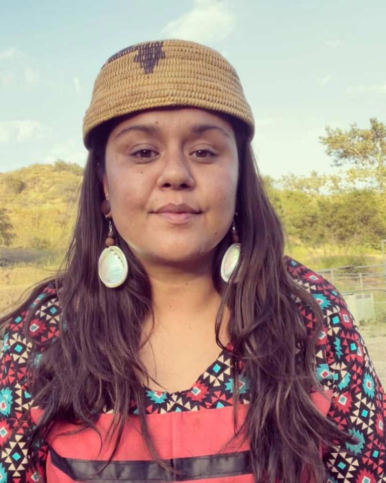 A woman with long brown hair wears a brown woven hat, large oval earrings, and a colorful patterned top. She stands outdoors with trees and hills in the background under a clear sky.