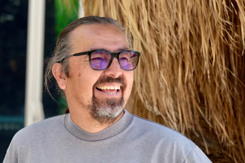 A man with long hair tied back, a goatee, and glasses with blue-tinted lenses, smiles while looking to the side. He is wearing a grey shirt and standing next to a large, straw-like plant.
